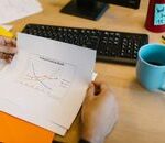 Close-up of hands reviewing a paper graph with a coffee cup and keyboard on a desk.